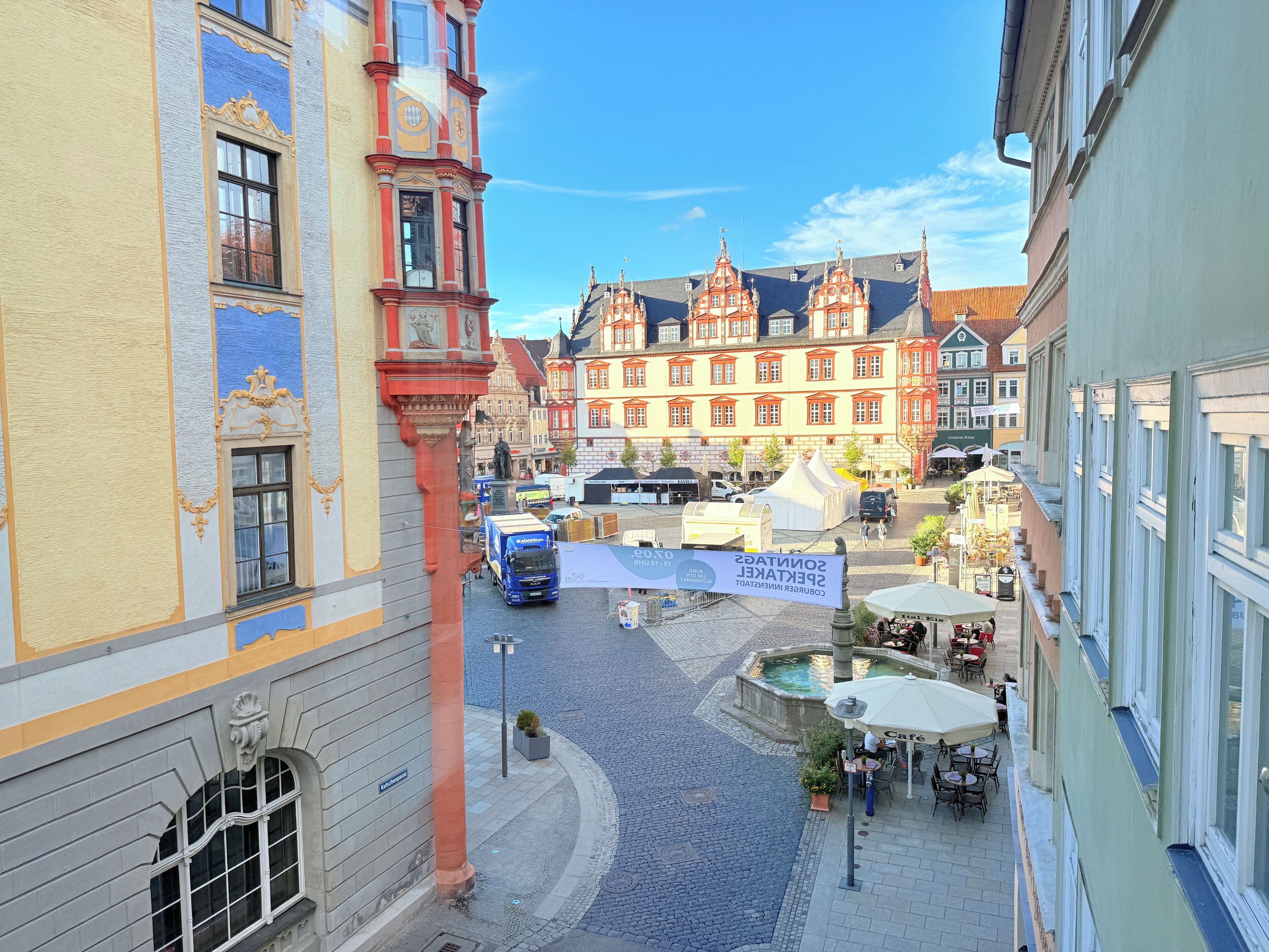 Ein Kopfsteinpflasterplatz mit historischen Gebäuden, einer Fontäne und Außenplätzen.