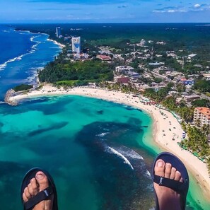A beach with clear turquoise waters, a sandy shore, and buildings in the background. - Sunset View 2BR Apartamento Juan Dolio Las Velas (Playa Juan Dolio San Pedro de Macoris)