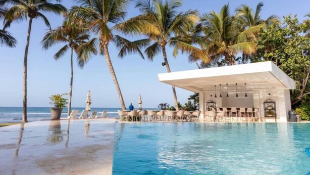 Een zwembadterras met ligbedden en parasols, palmbomen en uitzicht op de oceaan.