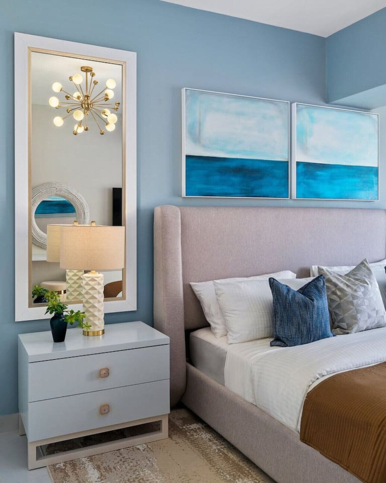 Une chambre à coucher avec un lit, une table de chevet avec une lampe, un miroir et deux tableaux abstraits accrochés au mur.