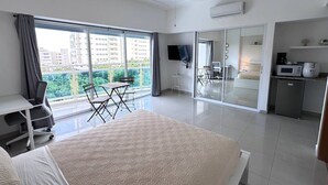 Ein modernes Hotelzimmer mit einem großen Bett, einem Schreibtisch, einem Sessel, einem Fernseher, einer Küchenzeile und einem Balkon mit Blick auf die Stadt.