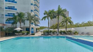 A swimming pool with lounge chairs and umbrellas, surrounded by palm trees and a modern building.