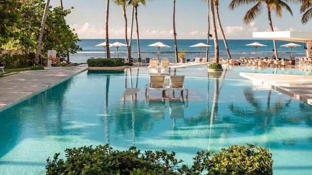 A pool with lounge chairs, palm trees, and a view of the ocean.