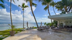 A poolside area with lounge chairs and tables, overlooking the ocean, with palm trees and a clear sky.