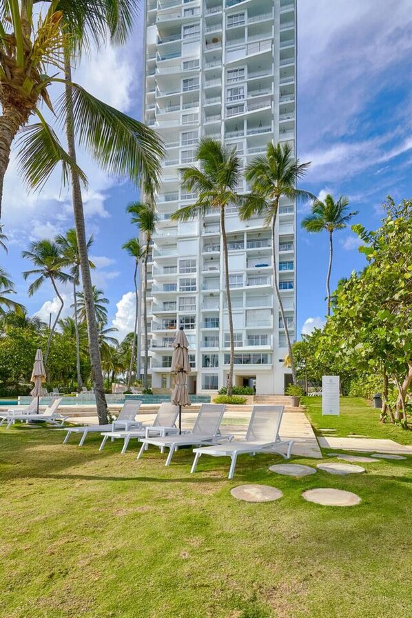 A high-rise hotel with a pool area, palm trees, and lounge chairs. - Sunset Caribbean Beachfront Gataway Aquarella Juan Dolio (Playa Juan Dolio)
