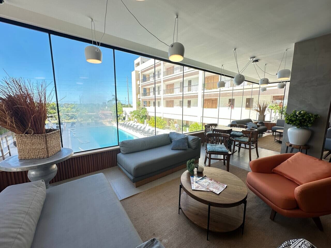 A modern living room with a large window overlooking a pool and a building. There are sofas, a coffee table, and hanging lights.