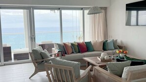 Een ruime woonkamer met een groot raam met uitzicht op zee, een bank met kleurrijke kussens en een salontafel met decoratieve objecten.
