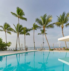A poolside area with lounge chairs and umbrellas, palm trees, and a view of the ocean.