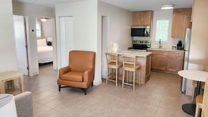 Une chambre d’hôtel moderne équipée d’un lit, d’un coin salon avec un fauteuil, d’une petite table et d’une kitchenette avec une cuisinière et un four à micro-ondes.