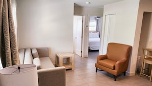 Une chambre d’hôtel avec un canapé, un fauteuil, une petite table et une lampe. On aperçoit un lit au fond de la pièce, et une porte donne accès à une autre chambre.