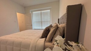 A bedroom with a large bed, a window with blinds, a door, and a mirror on the wall.