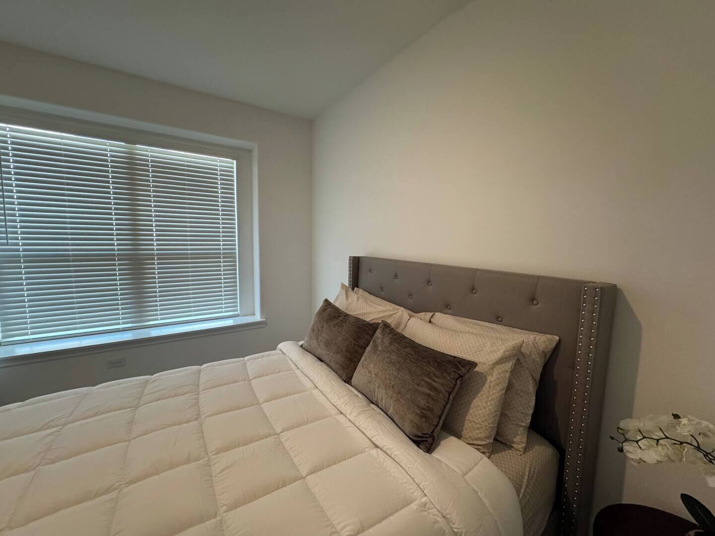 Quarto com cama grande, janela com persianas e mesa de cabeceira fixa na parede com um vaso de flores.