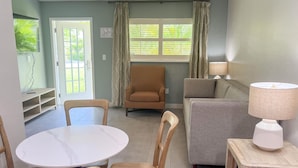 Uma sala de estar com uma mesa redonda, duas cadeiras, um sofá, um abajur e uma janela com cortinas.