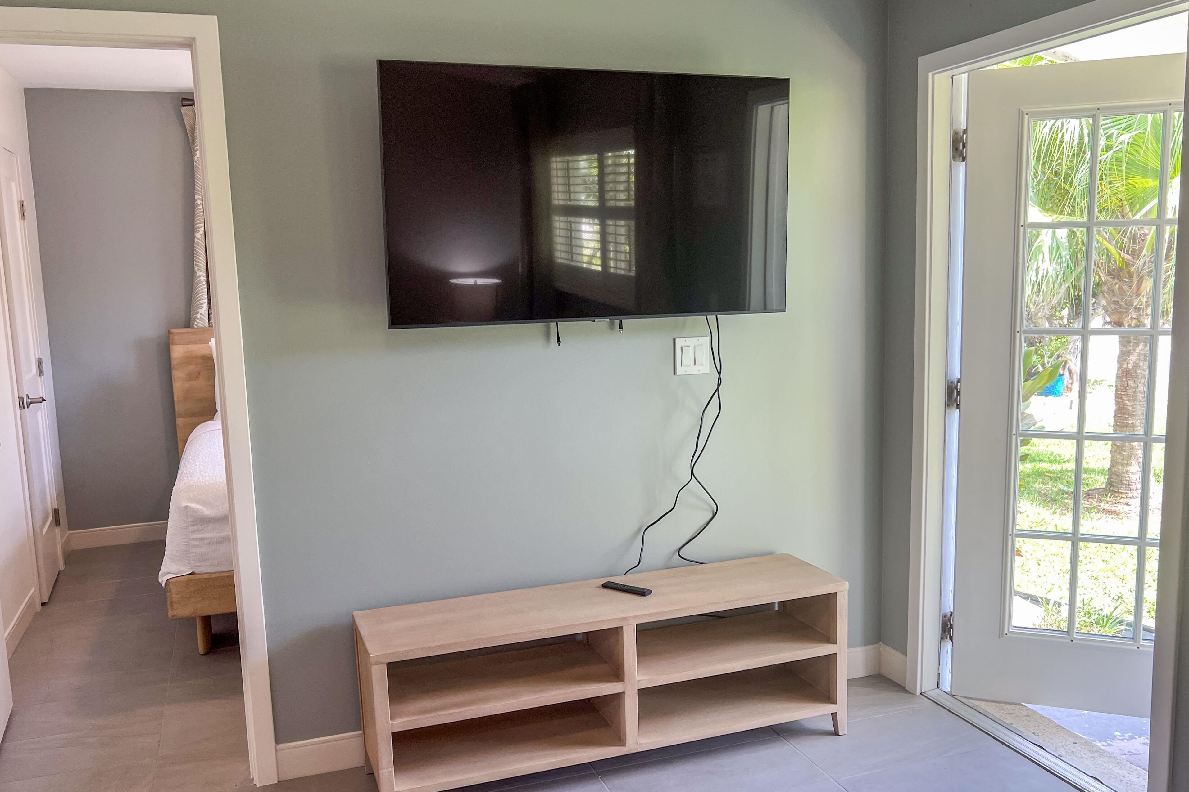 A room with a TV mounted on the wall, a wooden bench, a bed, and a door leading outside.
