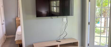 A room with a TV mounted on the wall, a wooden bench, a bed, and a door leading outside.