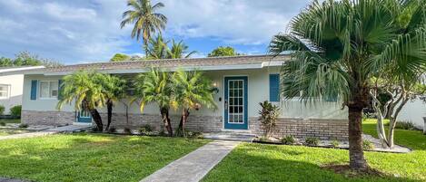 A single-story house with a brick facade, a blue door, and a small front yard with palm trees.