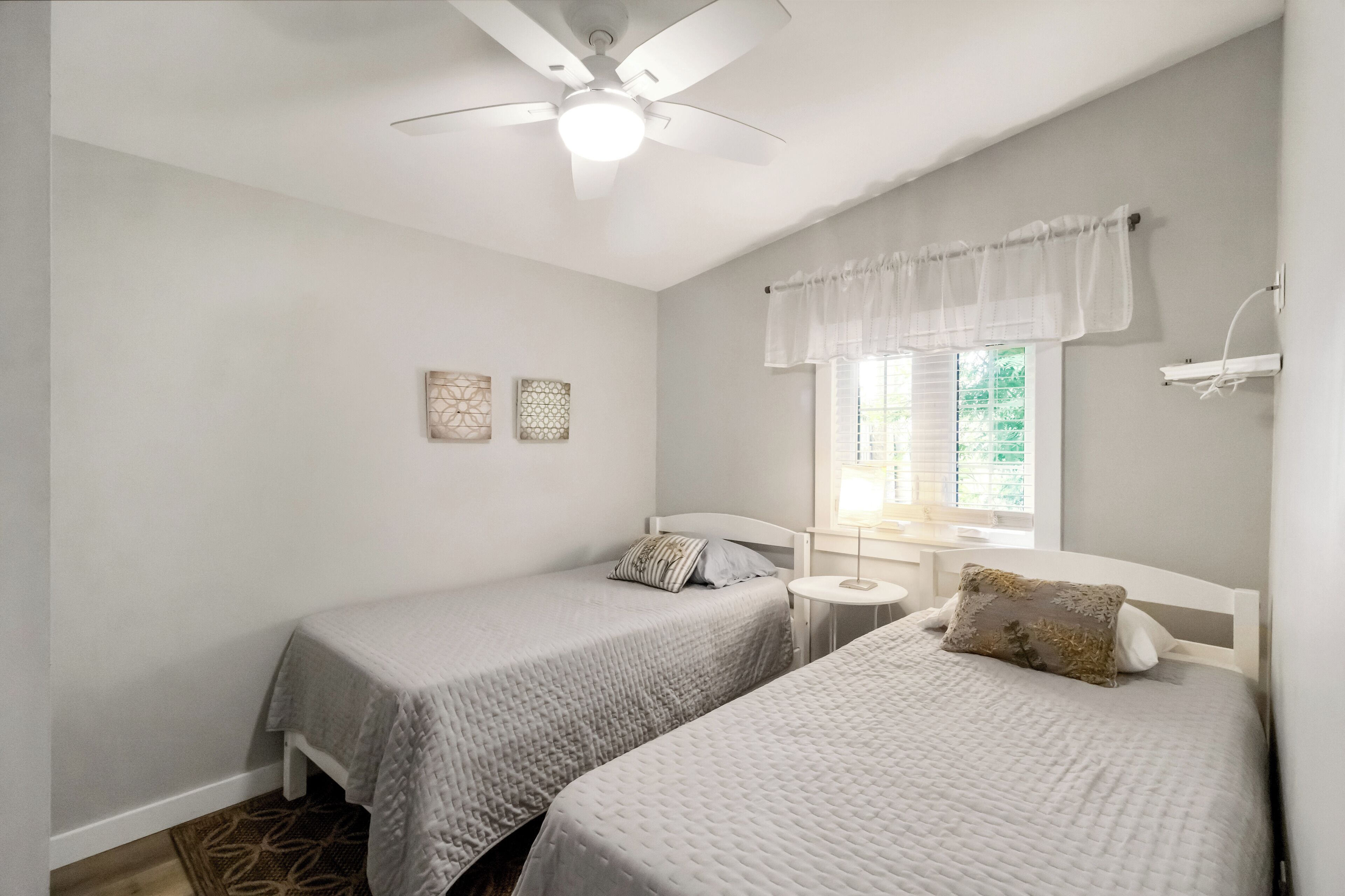A bedroom with two beds, a ceiling fan, a window with curtains, and a small table.