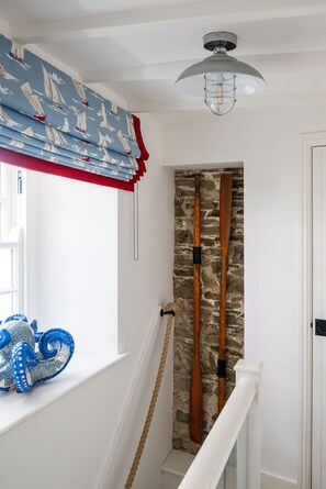 A staircase with a rope handrail, a wooden wall with two paddles, a window with a blue and white sailboat pattern curtain, and a white ceiling light fixture.