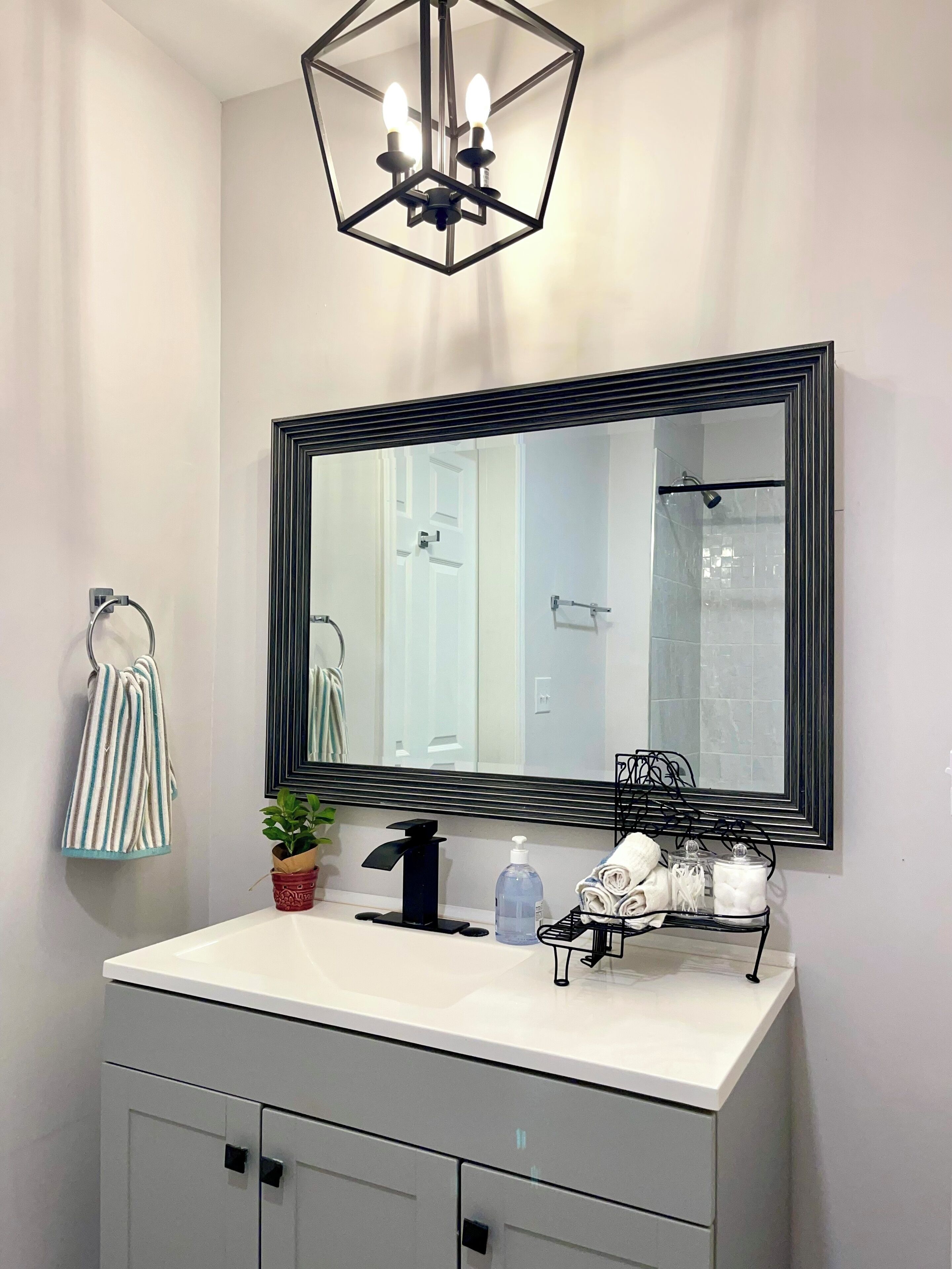 Une salle de bain avec un lavabo blanc, un grand miroir, un porte-serviettes, une plante en pot, un distributeur de savon et un autre porte-serviettes.