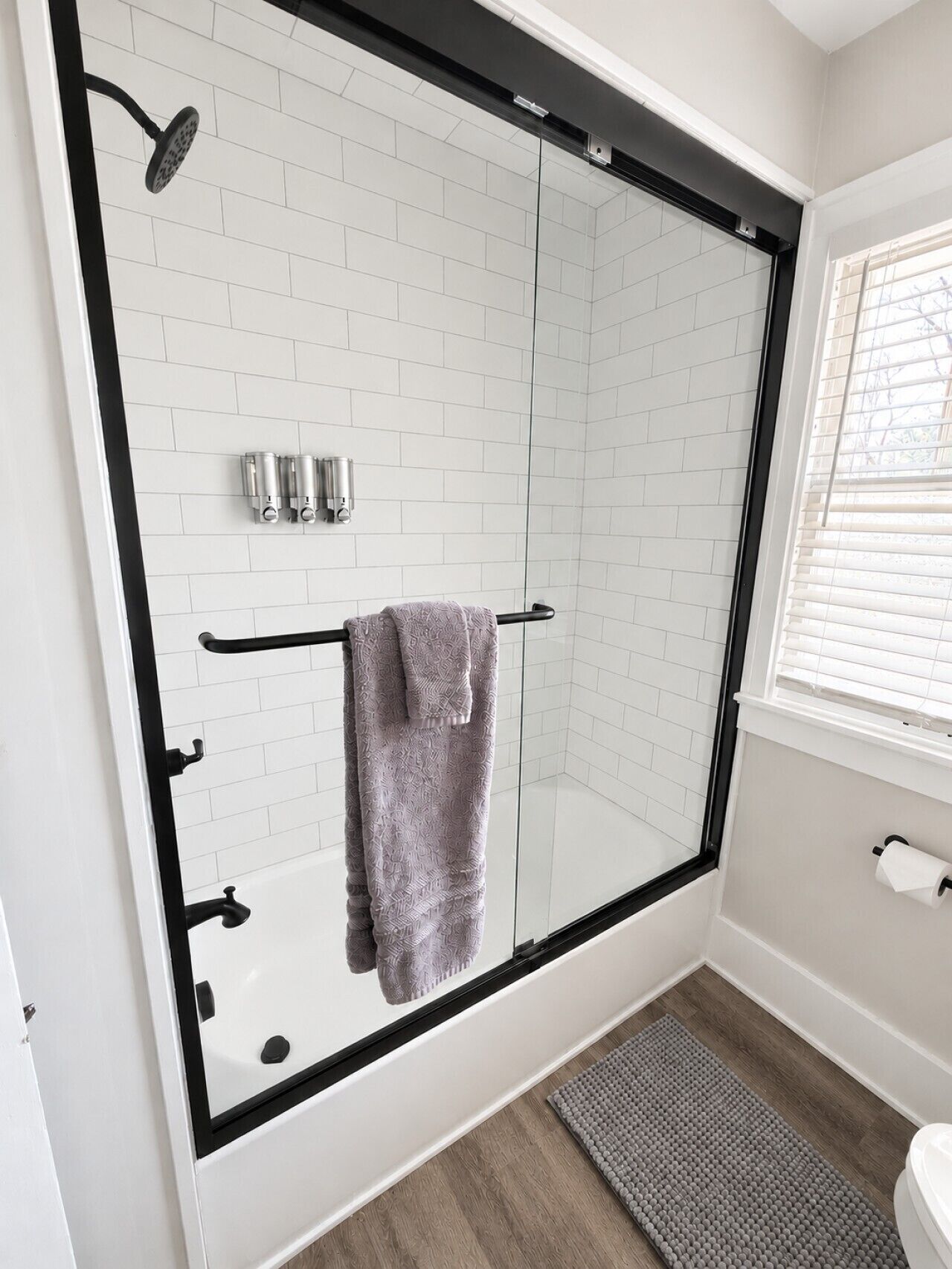 A modern bathroom with a glass-enclosed shower, white tiled walls, a towel rack, and a window with blinds.