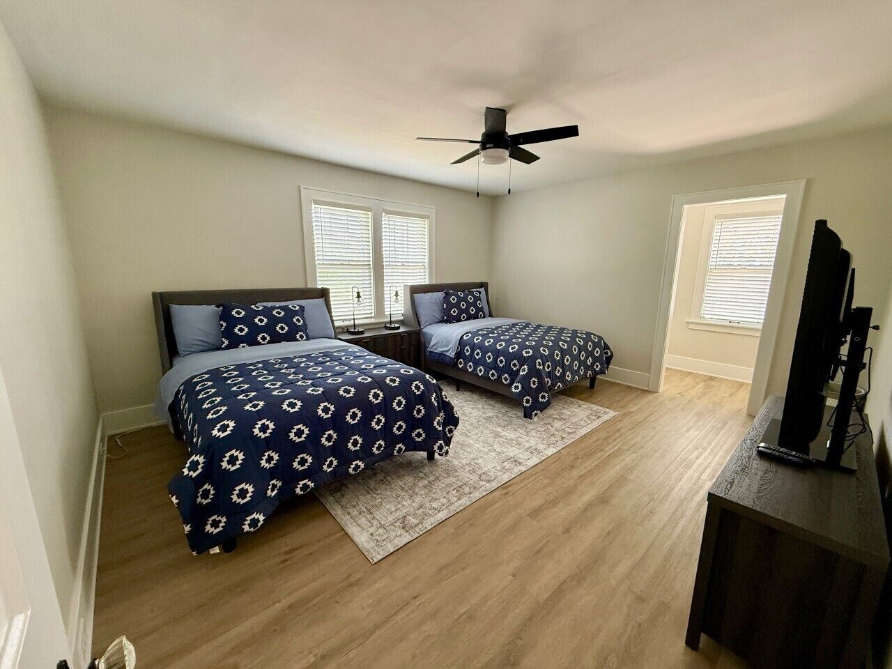 A bedroom with two beds, a ceiling fan, a TV stand, and a window with blinds.