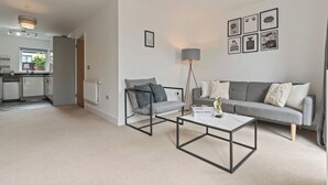En moderne stue med en grå sofa, et sofabord med sort ramme og en gulvlampe i grå. Et køkken ses i baggrunden.