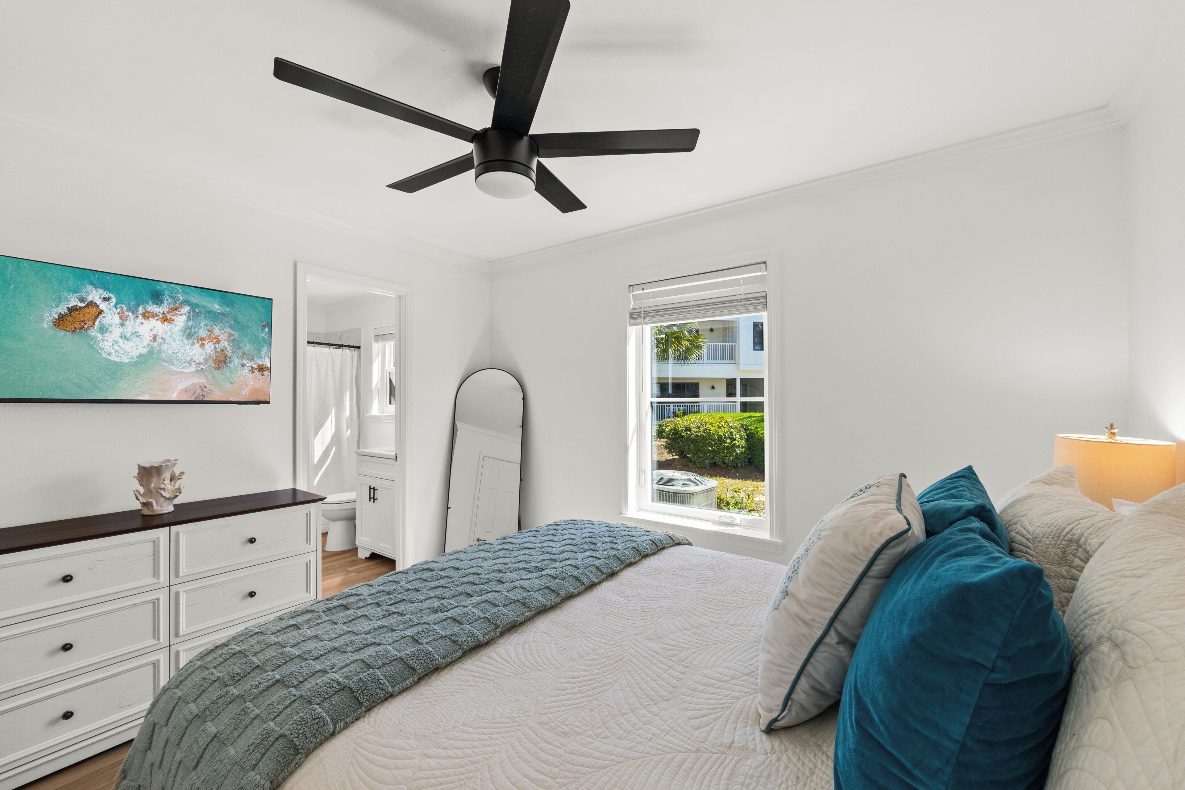 A bedroom with a bed, a ceiling fan, a window with blinds, a painting on the wall, and a dresser.