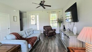 Una sala de estar con un sofá seccional, un sillón de cuero, una mesa de centro de madera, un televisor montado en la pared y un ventilador de techo.