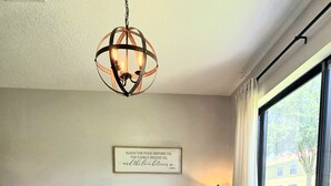 Comedor con mesa de madera dispuesta para seis personas, rodeada de sillas, una lámpara colgante y una cita inspiradora enmarcada en la pared.