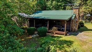 A rustic stone cabin with a green roof, surrounded by lush greenery and a wooden deck. - Almost Heaven – Porch Swing Mountain Getaway (Franklin)
