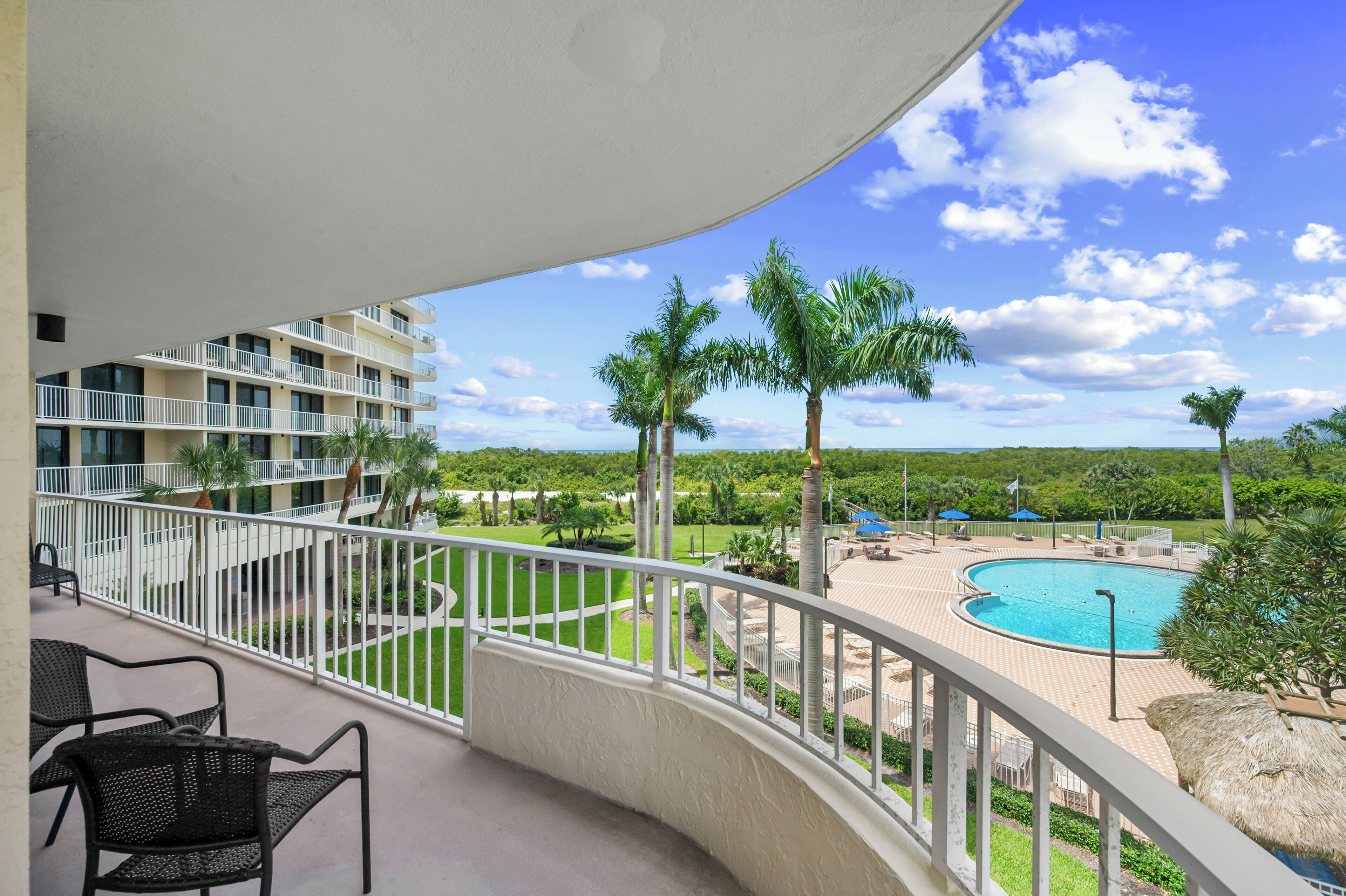A balcony with a view of a swimming pool, palm trees, and a multi-story building.