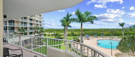 A balcony with a view of a swimming pool, palm trees, and a multi-story building.