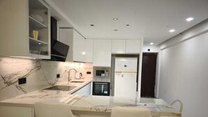 A modern kitchen with a marble countertop, a built-in oven, and a refrigerator. There are two white bar stools at the island. - Airbhouse Prívate Rooms & Triana (Seville)