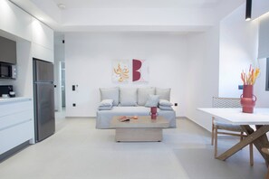 A modern kitchen with a stainless steel refrigerator, a microwave, and a coffee machine. There is a light-colored sofa with cushions and a coffee table in front. A dining table with a vase of flowers is visible. - Achillion Suites (Lefkada)