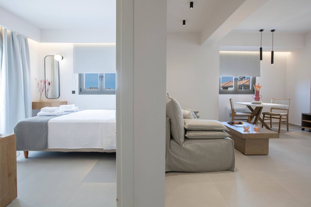 A modern hotel room with a bed, a seating area with a table and chairs, and a view of the outside.