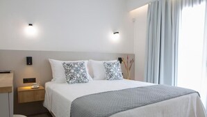 A modern hotel room with a large bed, a bedside table with a lamp, and a view of the outdoors through the window. - Achillion Suites (Lefkada)