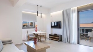 A modern living room with a sofa, coffee table, dining table, and chairs. There is a TV mounted on the wall and a sliding glass door leading to an outdoor area. - Achillion Suites (Lefkada)