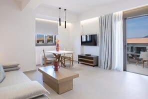 A modern living room with a sofa, coffee table, dining table, and chairs. There is a TV mounted on the wall and a sliding glass door leading to an outdoor area. - Achillion Suites (Lefkada)