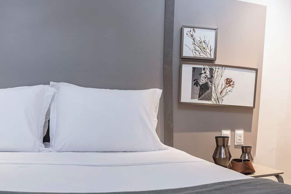 Ein ordentlich bezogenes Bett mit weißen Leinen und Kissen, ein Nachttisch mit Lampe sowie zwei gerahmte Bilder an der Wand.