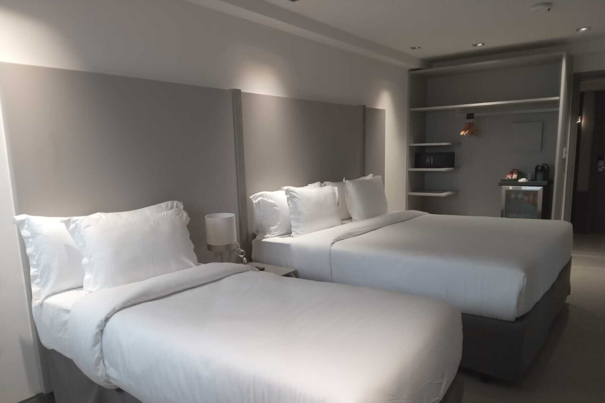 Ein Hotelzimmer mit zwei Betten, einem Nachttisch, einer Lampe, einem Kleiderschrank und einer kleinen Küchenzeile.