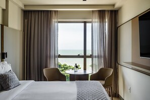 Une chambre d’hôtel avec un lit, deux chaises, une petite table, un téléviseur et une vue sur la mer grâce à de grandes fenêtres.