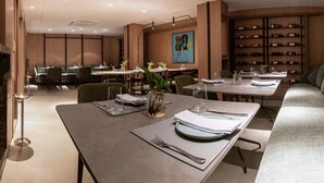 Un comedor moderno con varias mesas, sillas y un sofá. Hay estanterías con libros y zapatos, y un cuadro en la pared.