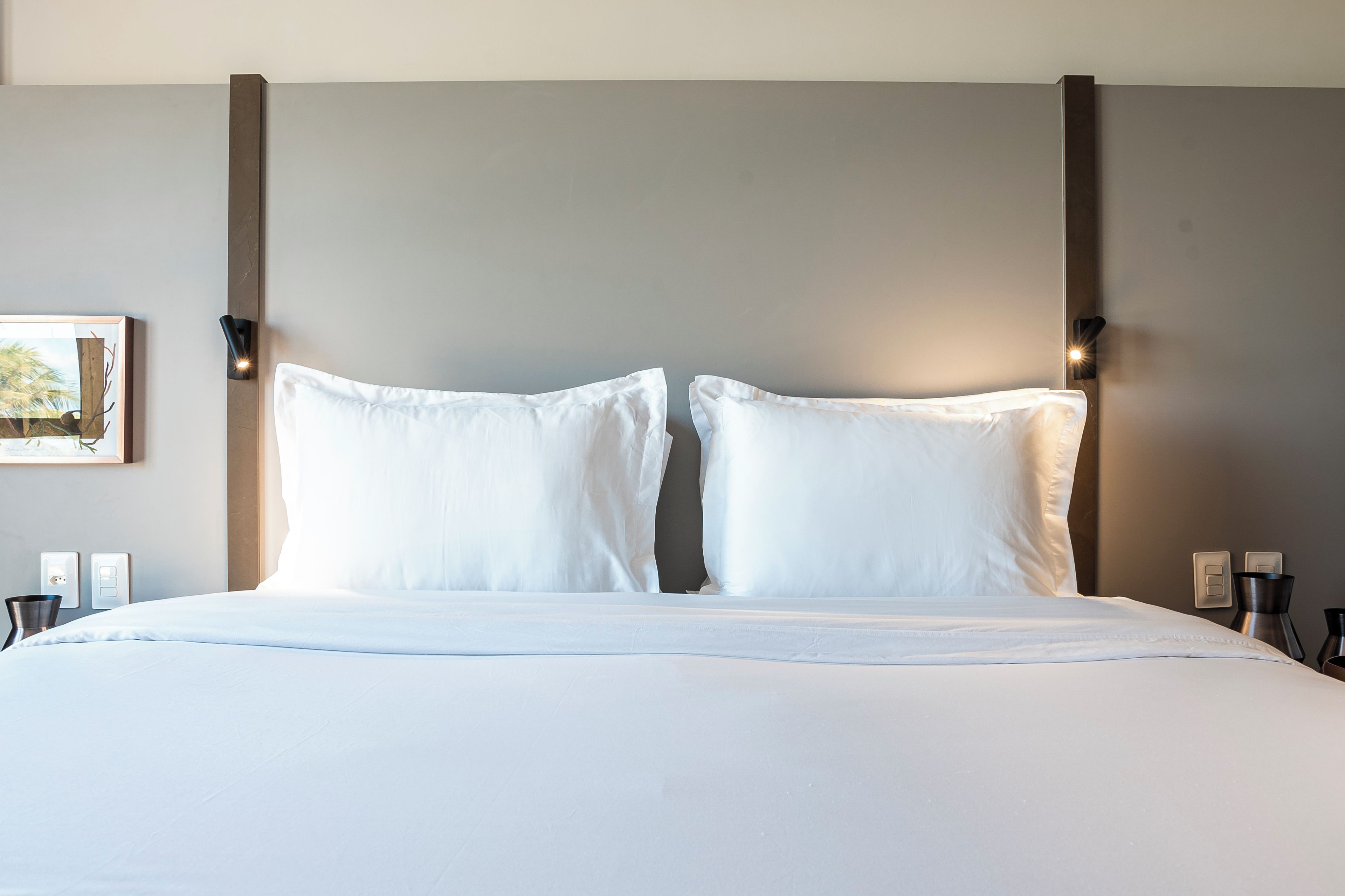 Una cama bien tendida con sábanas blancas, una lámpara de pared y un cuadro en la pared.