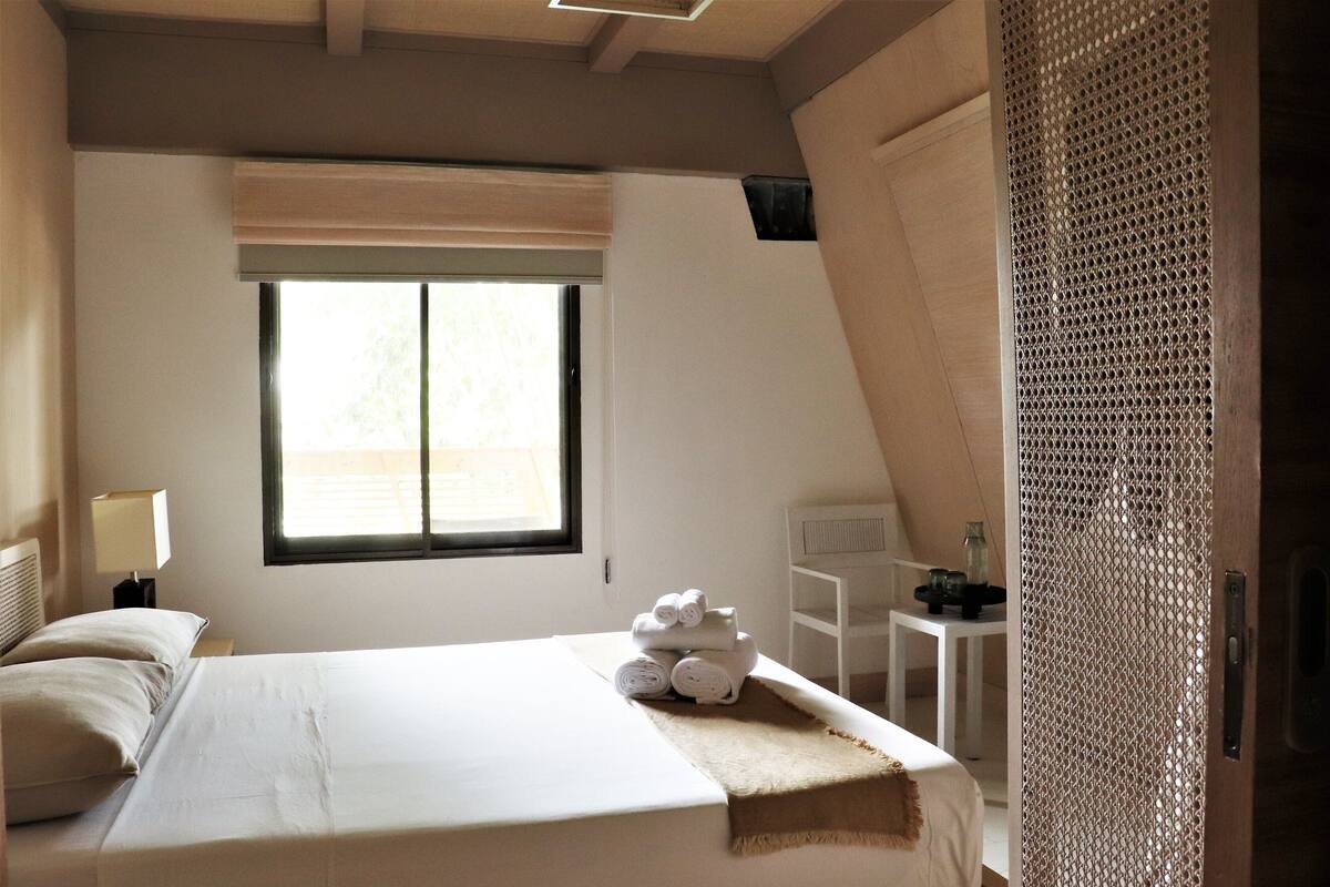Ein Schlafzimmer mit Bett, Fenster, Nachttisch und einem kleinen Tisch mit Lampe.