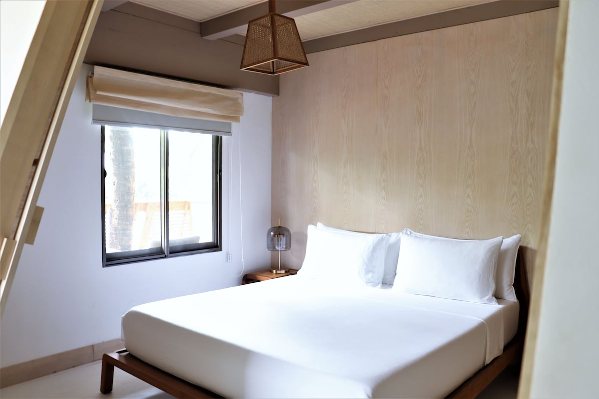 A bedroom with a bed, a window with a blind, a nightstand with a lamp, and a sloped ceiling.