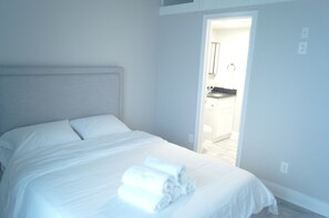 Une chambre à coucher avec un lit, un lit bien fait avec des serviettes à portée de main, une porte donnant sur une salle de bain et une cuisine visible.