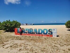 A beach with a large sign reading 'MARADOS' sponsored by KFC, a clear blue sky, and the ocean in the background.