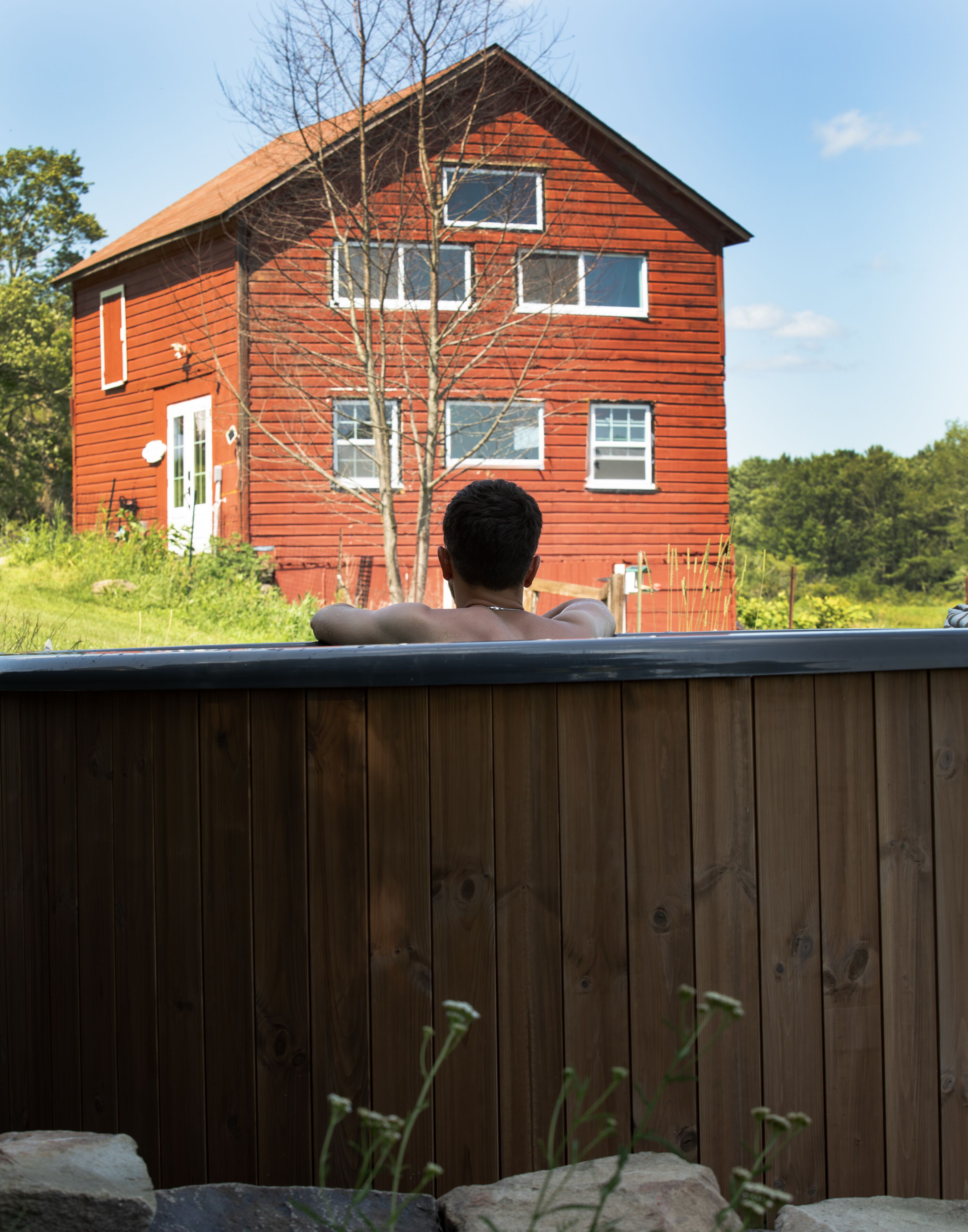 Une maison en bois rouge, dotée de nombreuses fenêtres et d’une porte blanche, vue de derrière une clôture en bois.