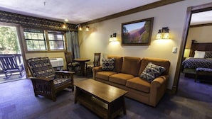 Un salon confortable avec un canapé, un fauteuil et une table basse. Il y a des fenêtres avec des rideaux, un tableau au mur et une porte qui donne sur une autre pièce.