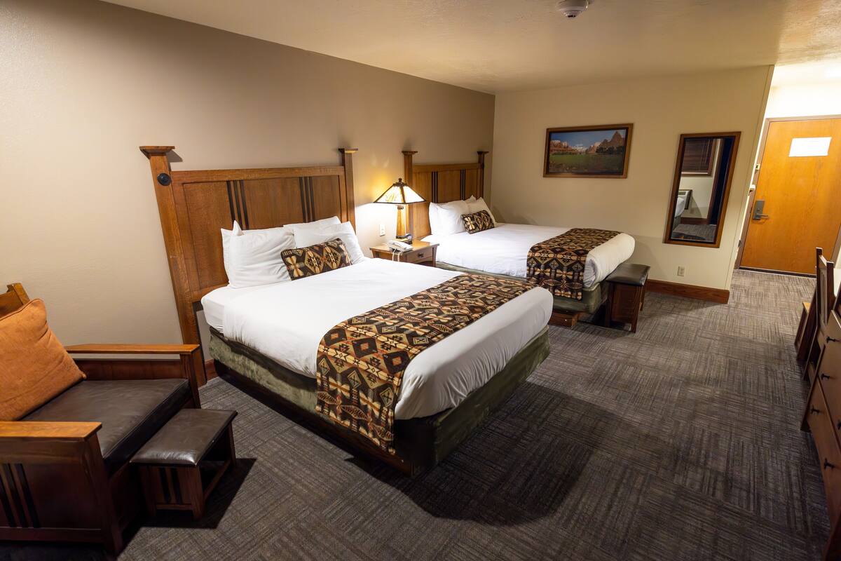 Habitación de hotel con dos camas, cabecero de madera, mesita de noche con lámpara, un banco y cómoda de madera.